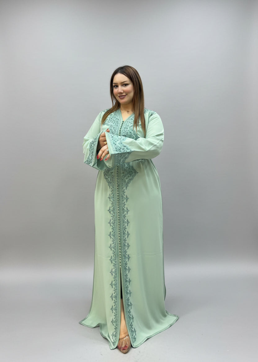 A woman wearing a light mint green long kaftan dress with blue embroidery.