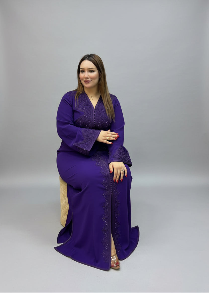 A woman wearing a long-sleeved purple kaftan with embroidered details sits.
