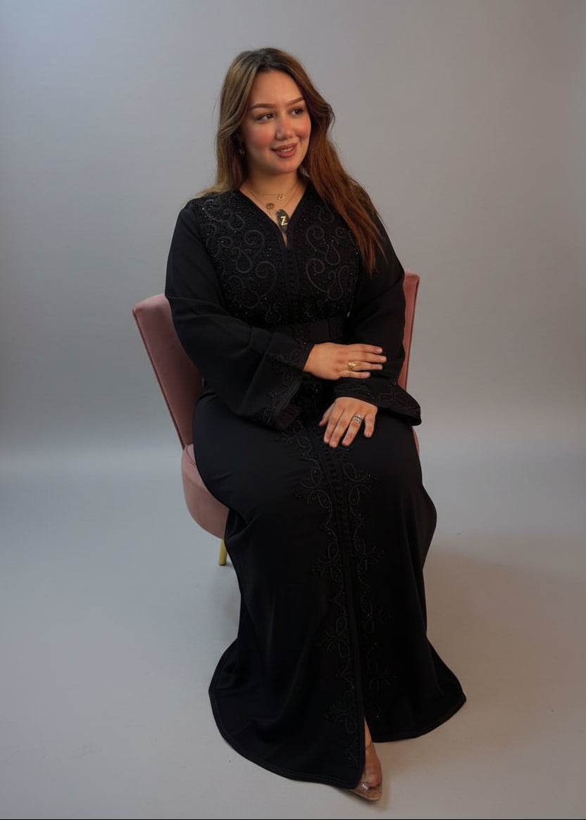 A woman wearing a long black embroidered moroccan caftan sits in a pink chair .