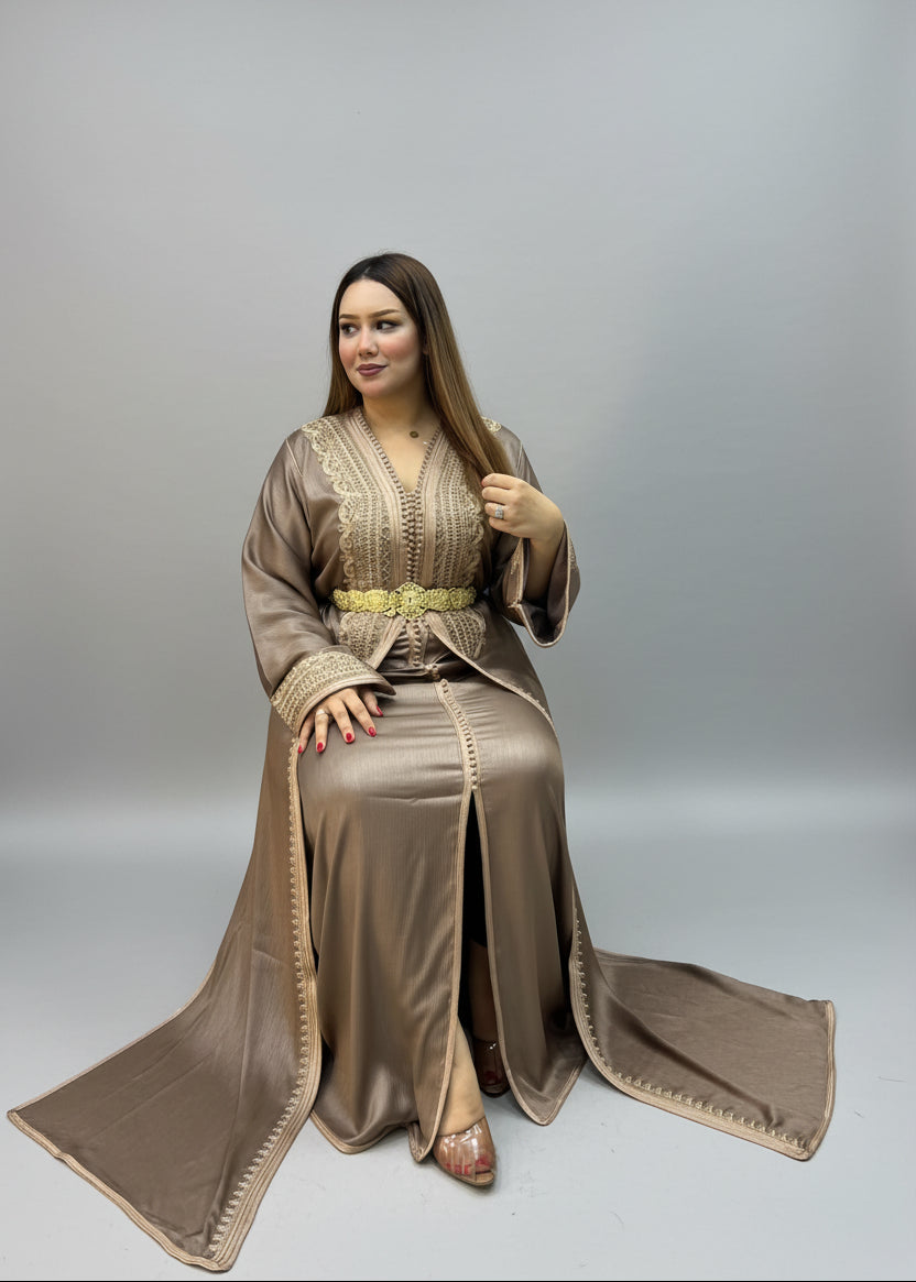 A woman wearing a traditional long brown kaftan with gold embroidery and a matching skirt sits .