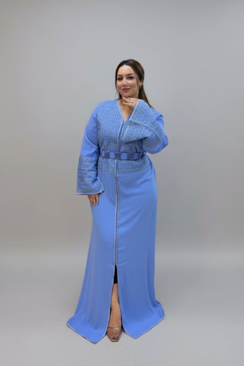A woman modeling a light blue traditional long kaftan with a textured embroidered yoke and matching belt, standing against a plain gray studio background.
