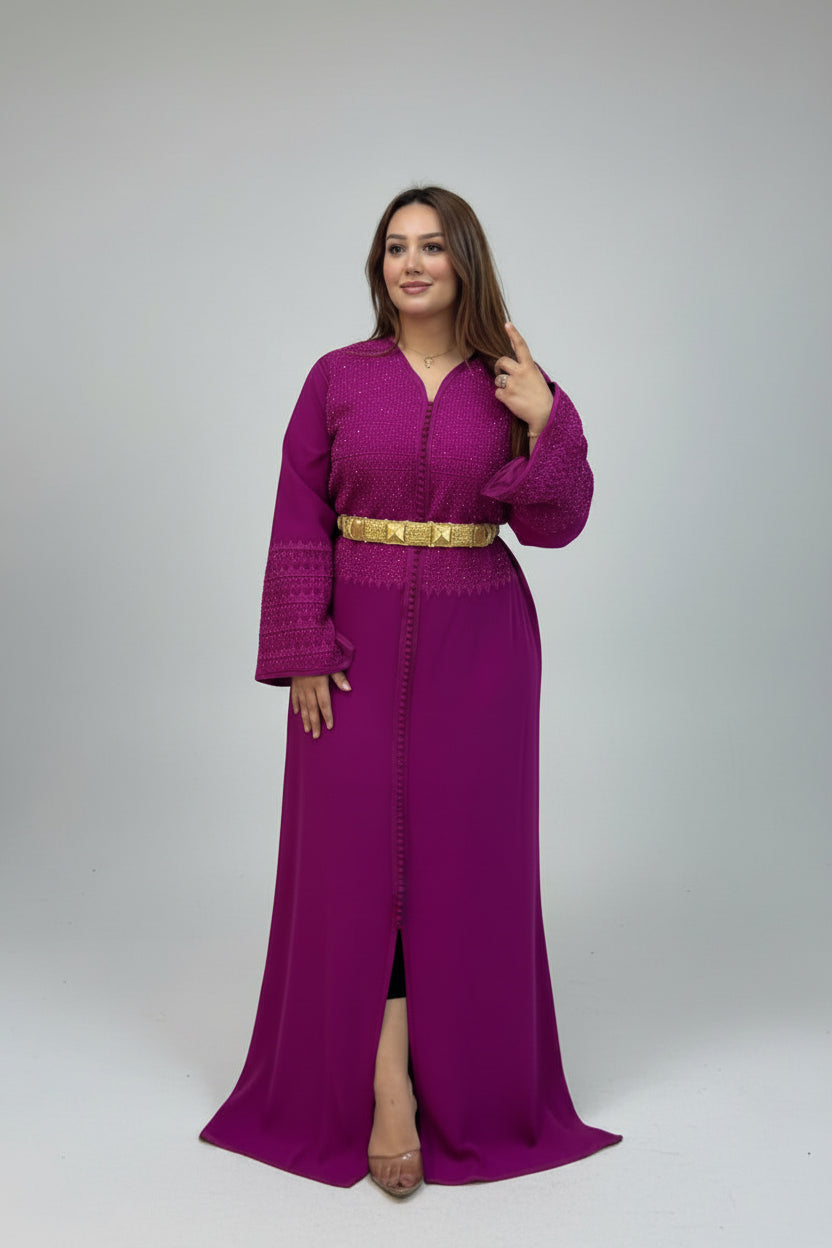 A woman modeling a long magenta-purple moroccan kaftan with a gold belt, standing against a light gray studio background.