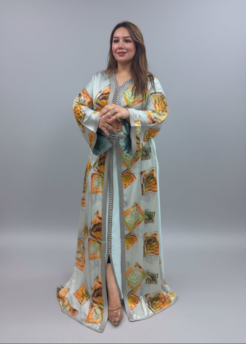A woman modeling a long, light sage green kaftan dress with orange and yellow geometric patterns.
