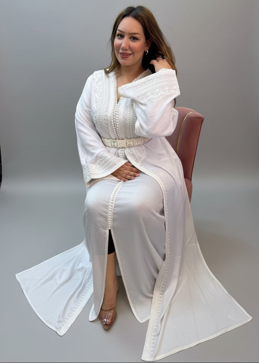 A woman wearing a white embroidered long kaftan sits on a chair .