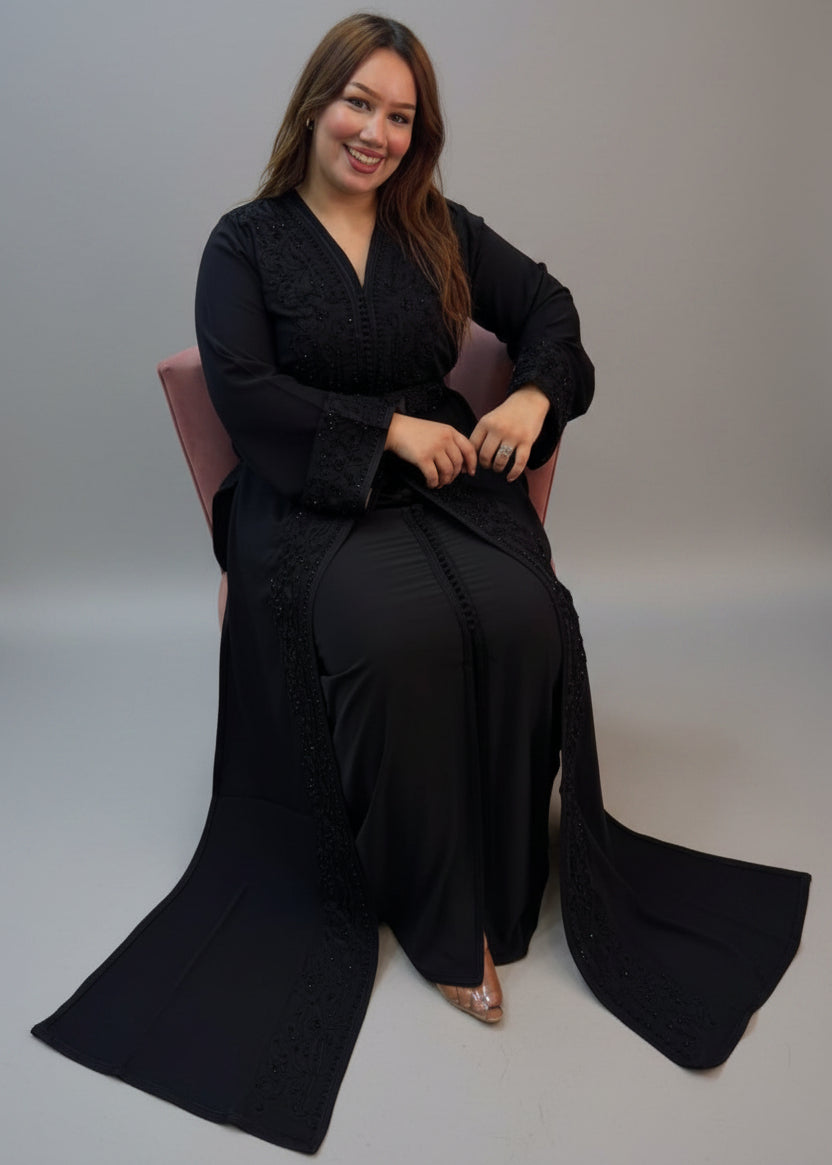 A woman wearing a long black embroidered kaftan and matching skirt sits in a chair.
