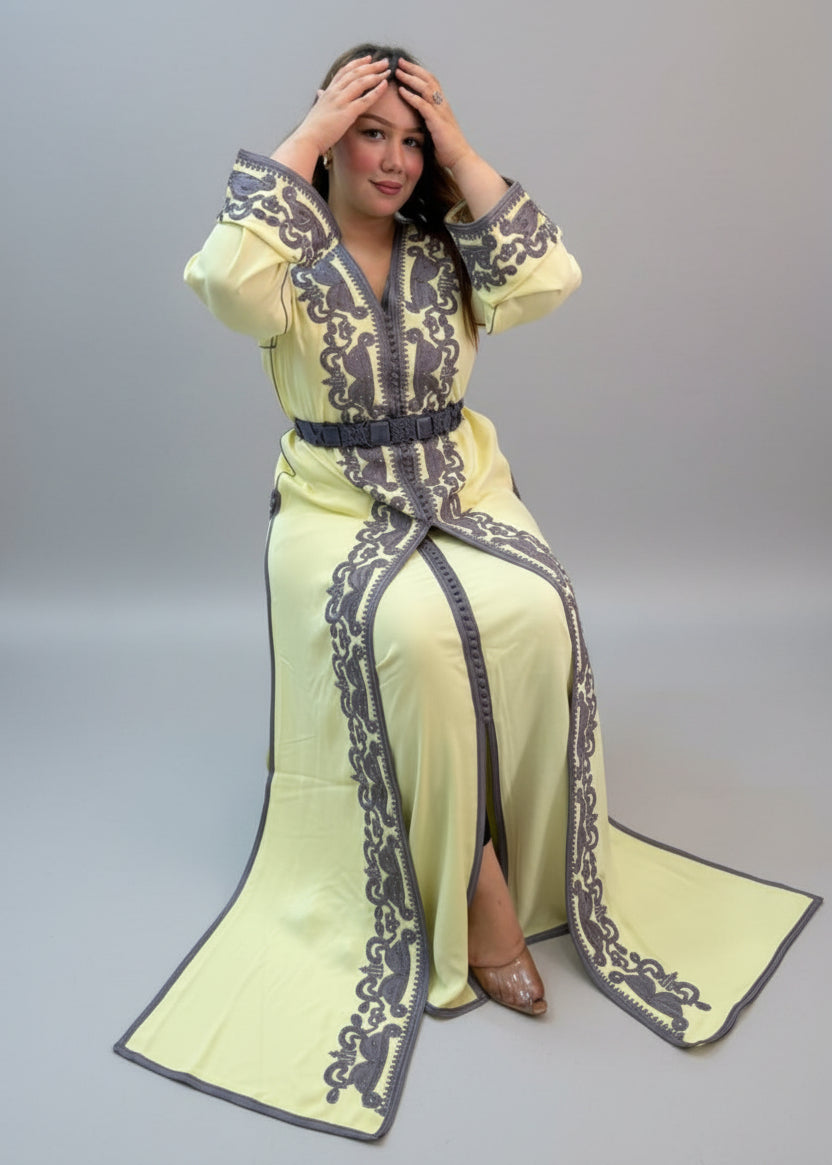 A woman wearing a pale yellow traditional long kaftan with dark gray embroidered patterns and a matching belt.