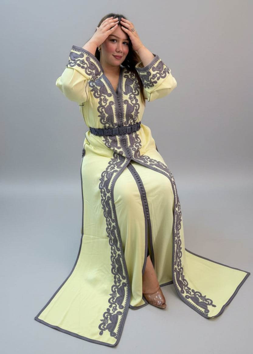 A woman wearing a pale yellow traditional long kaftan with dark gray embroidered patterns and a matching belt.