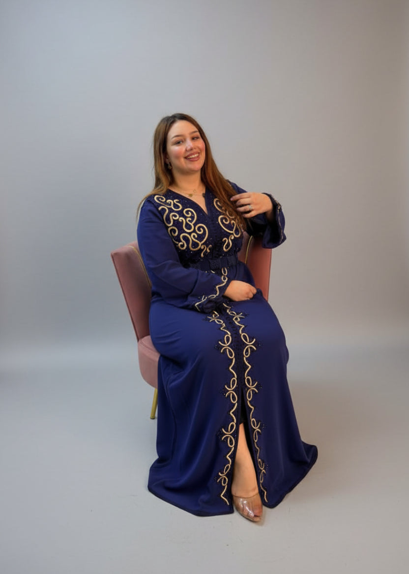 A woman wearing a navy blue long marocian kaftan with gold embroidery sits in a chair.