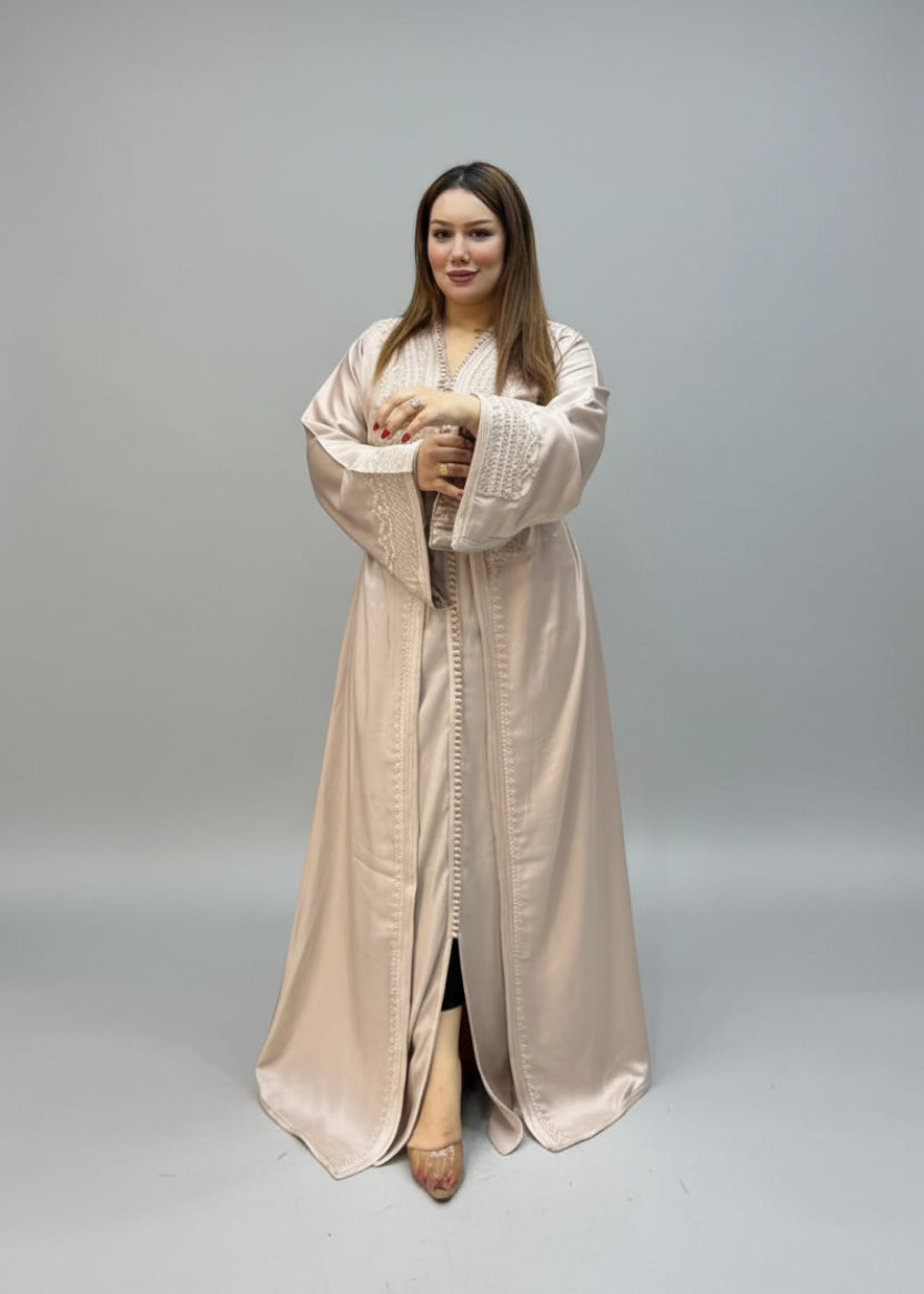 A woman wearing a long, beige satin kaftan with embroidered details .
