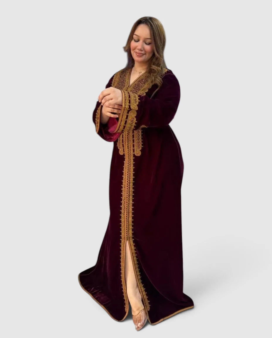A woman modeling a long, floor-length maroon velvet Kaftan with ornate gold embroidery and a high slit, posed against a light gray studio background.