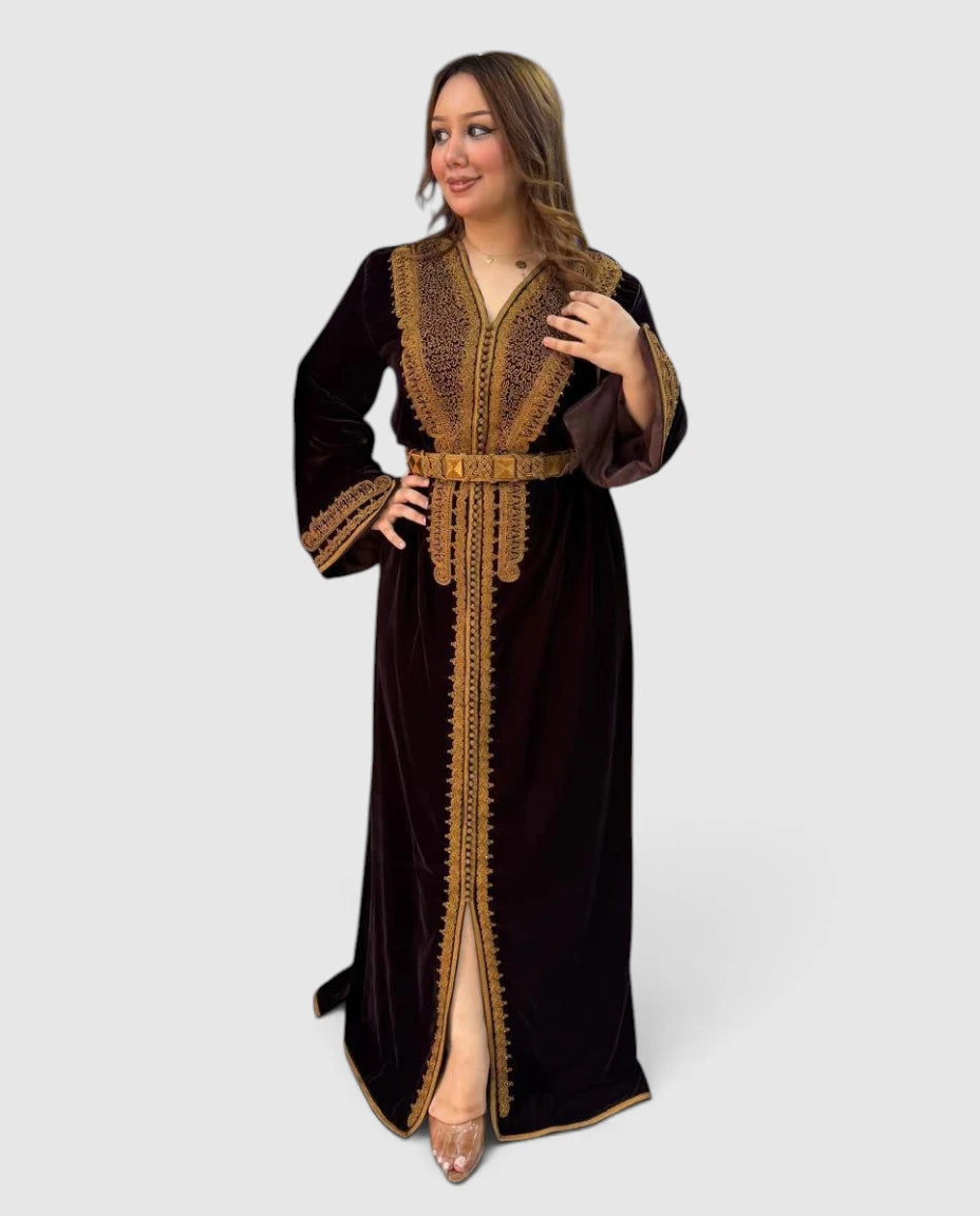A woman modeling a long dark brown velvet caftan with ornate gold embroidery and a matching belt, set against a solid light gray background.