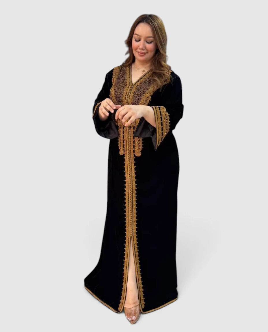A woman modeling a long black caftan dress with ornate gold embroidery and a high front slit, photographed against a light gray studio background.