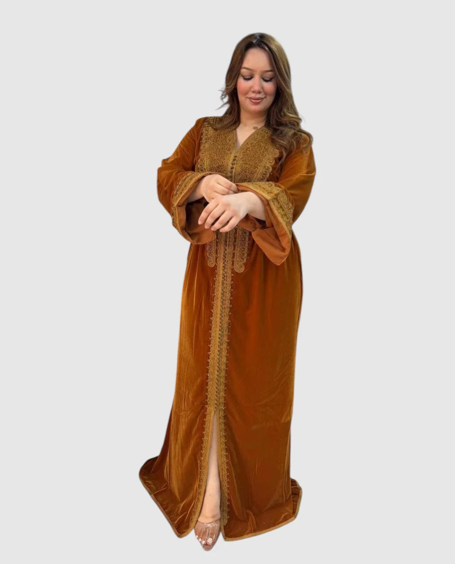 A woman modeling a long, flowing kaftan dress in a warm camel brown velvet fabric with gold embroidery and lace trim, set against a solid light gray background.