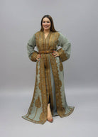 A woman modeling a sage green and gold embroidered kaftan dress .