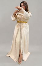 A woman modeling a long cream-colored Moroccan caftan (djellaba) dress with gold embroidery, a gold belt, and clear high-heeled sandals, posed against a solid gray studio background.