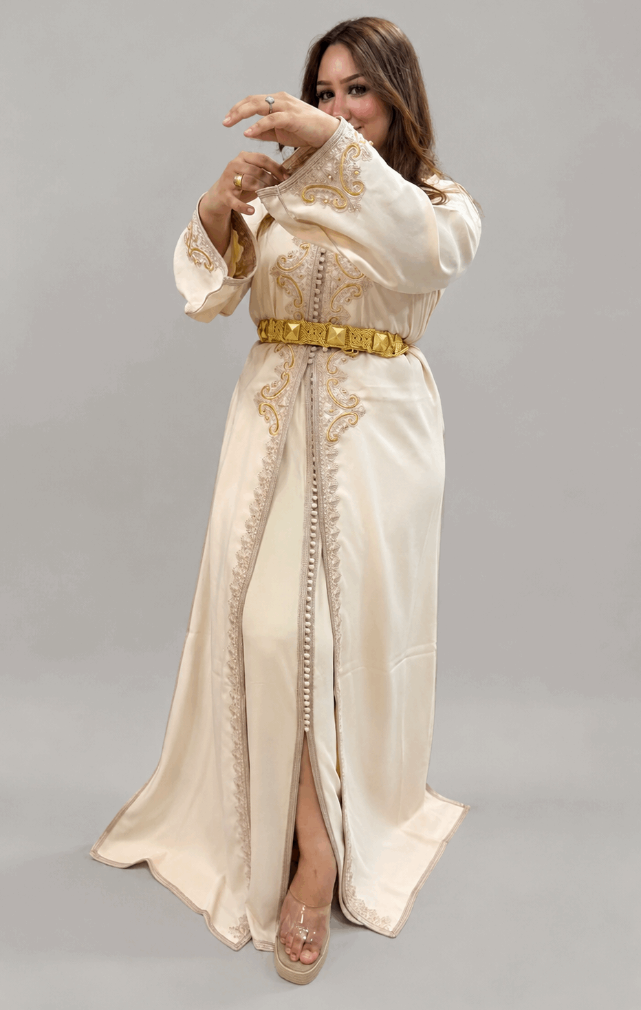 A woman modeling a long cream-colored Moroccan caftan (djellaba) dress with gold embroidery, a gold belt, and clear high-heeled sandals, posed against a solid gray studio background.