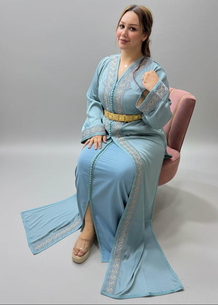 A woman wearing a light blue traditional long kaftan with silver embroidery and a gold belt, sitting on a pink chair inside a Caftan store.