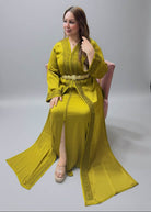 A woman wearing a long olive green kaftan dress with gold embroidery and a matching belt, posing in a Caftan store.
