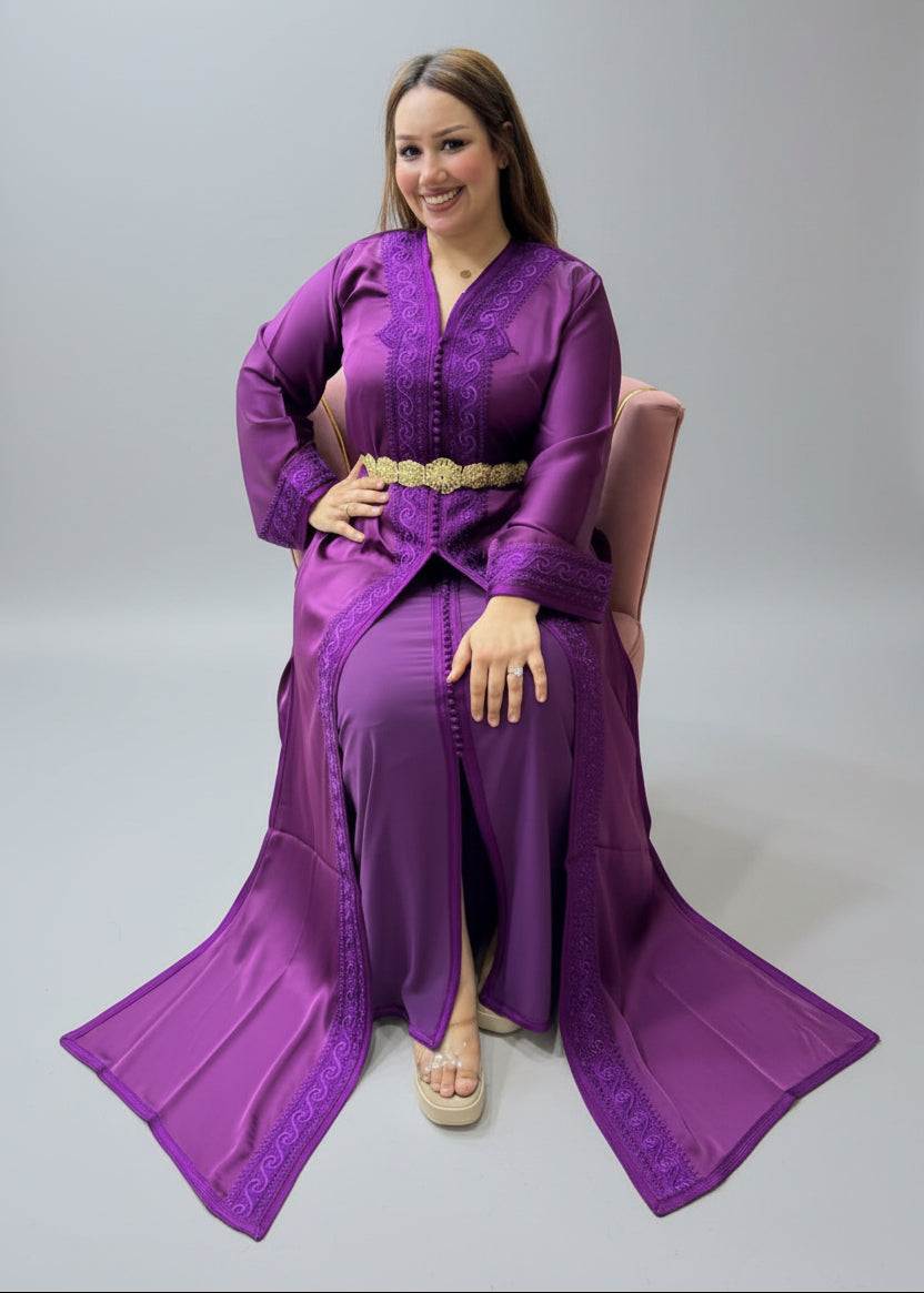 A woman wearing a long purple traditional kaftan or takchita with a gold belt, sitting in a chair inside a Caftan store.