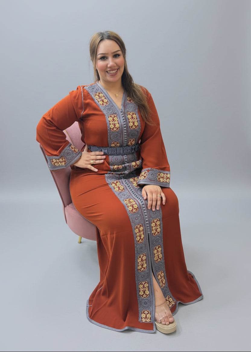 A woman wearing a rust-orange traditional long Caftan with gray and gold embroidered borders, seated in a chair inside a kaftan  store.