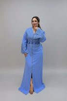 A woman modeling a light blue traditional long kaftan with a textured embroidered yoke and matching belt, standing against a plain gray studio background.