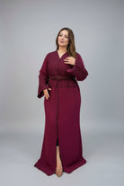A woman modeling a long, floor-length maroon kaftan with long sleeves, a matching belt, and a front slit, photographed against a neutral gray studio background.