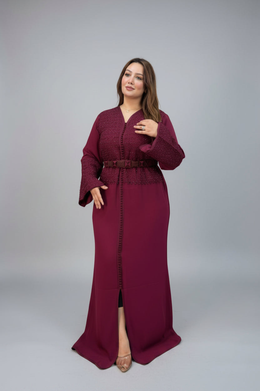 A woman modeling a long, floor-length maroon kaftan with long sleeves, a matching belt, and a front slit, photographed against a neutral gray studio background.
