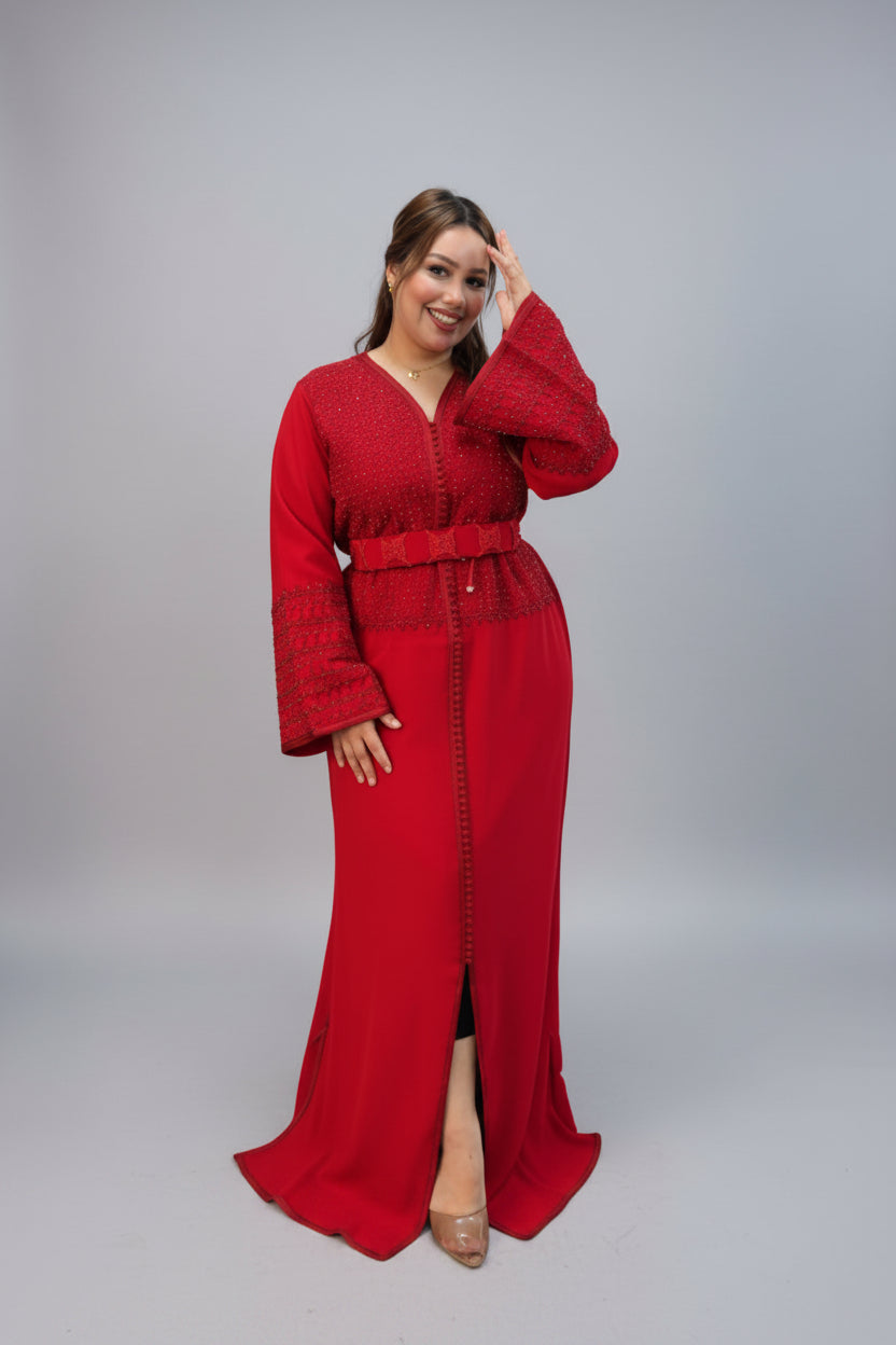 A woman modeling a long-sleeved red kaftan  with a textured lace-like bodice, matching waist tie, and front slit, standing against a plain gray studio background.