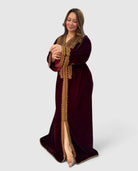 A woman modeling a long, floor-length maroon velvet Kaftan with ornate gold embroidery and a high slit, posed against a light gray studio background.