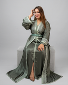 A woman wearing a long sage green Moroccan caftan with intricate silver embroidery, sitting on a light gray block against a neutral gray background.