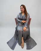 A woman wearing a long, flowing gray kaftan with embroidered details sits on a pink chair against a light gray studio background.
