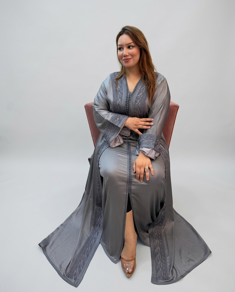 A woman wearing a long, flowing gray kaftan with embroidered details sits on a pink chair against a light gray studio background.