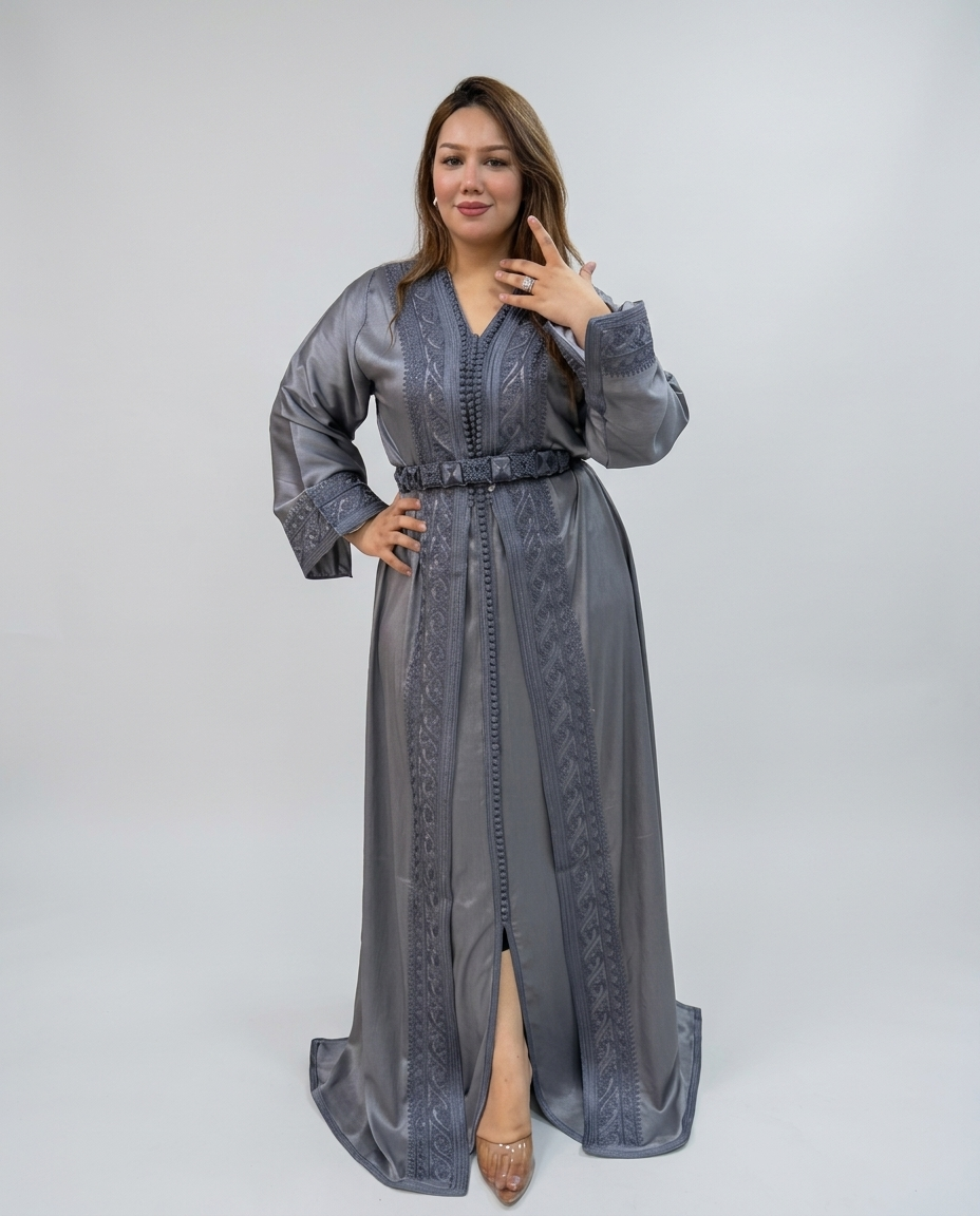 A woman modeling a long gray kaftan dress with intricate embroidery, a matching belt, and a front slit, photographed against a light gray studio background.