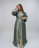 A woman modeling a long sage green kaftan robe with intricate light green embroidery, a matching belt, and a front slit, photographed against a neutral light gray studio background.