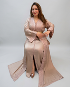 A woman models a long, flowing kaftan dress in a dusty rose pink color with matching embroidered lace details. She is seated against a plain light gray studio background.