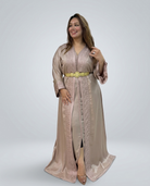 A woman modeling a long beige satin caftan with intricate embroidery, a gold belt, and matching embroidered sleeves, set against a plain light gray studio background.