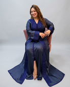 A woman wearing a long navy blue satin caftan with intricate embroidery, seated on a pink chair against a light gray studio background.