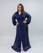 A woman modeling a long navy blue satin caftan with intricate embroidery and a matching waist belt, set against a plain light gray studio background.
