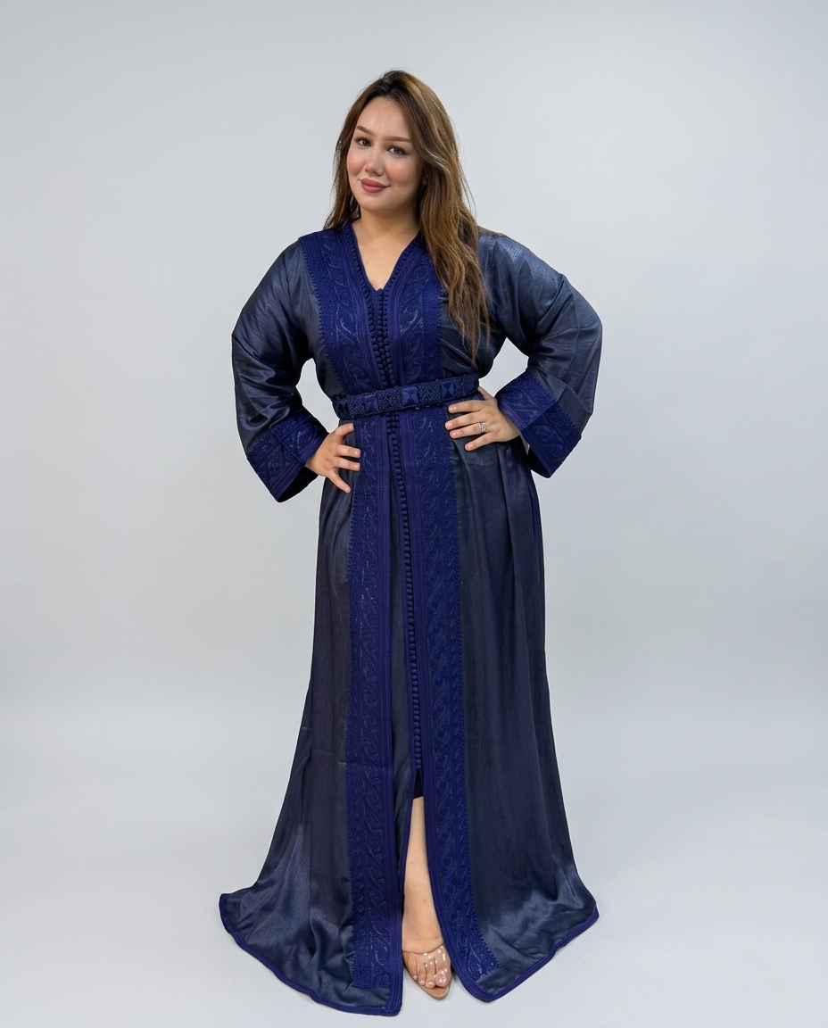 A woman modeling a long navy blue satin caftan with intricate embroidery and a matching waist belt, set against a plain light gray studio background.