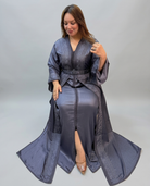 A woman wearing a long, floor-length gray kaftan with wide sleeves, a V-neck, and a matching waist tie, posed against a plain light gray studio background.