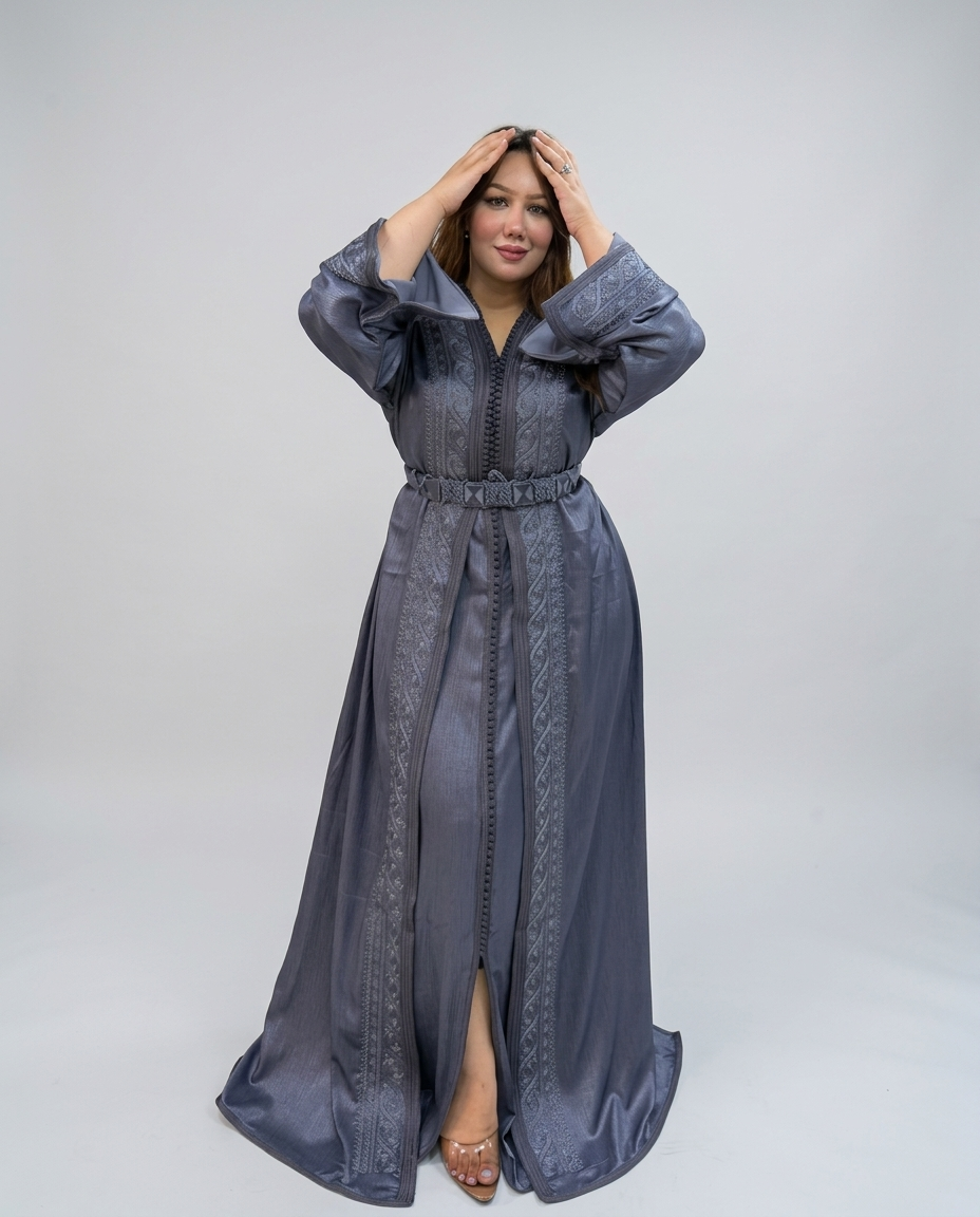 A woman modeling a long, floor-length gray abaya dress with a matching waist tie and embroidered details, standing against a light gray studio background.