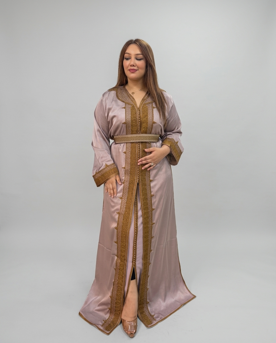 A woman modeling a long satin-like kaftan in a dusty pink color with prominent gold embroidery and trim, standing against a neutral light gray studio background.