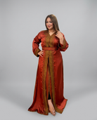A woman modeling a long rust-orange satin caftan with ornate gold embroidery and a matching belt, photographed against a neutral gray studio background.