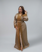 A woman modeling a long beige satin Caftan with ornate gold embroidery and a matching belt, photographed against a neutral gray studio background.