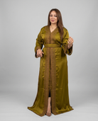 A woman modeling a long olive green satin kaftan with gold embroidered borders and a matching belt, standing against a light gray studio background.