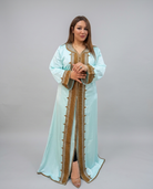 A woman modeling a long, flowing light blue kaftan dress with ornate gold embroidery and trim, standing against a neutral gray studio background.