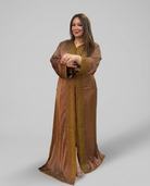 A woman modeling a long brown kaftan with gold embroidered trim and a matching brown headscarf, standing against a solid gray studio background.