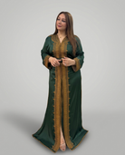 A woman modeling a dark green kaftan dress with ornate gold embroidery and trim, standing against a solid gray studio background.