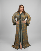 A woman modeling a long, loose caftan-style dress with wide sleeves, a V-neckline, and a front slit, featuring an olive green base with a metallic gold abstract pattern and dark trim. She is standing against a neutral light gray studio background.