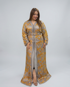 A woman modeling a long, floor-length kaftan dress with a gold and gray geometric pattern, standing against a light gray studio background.